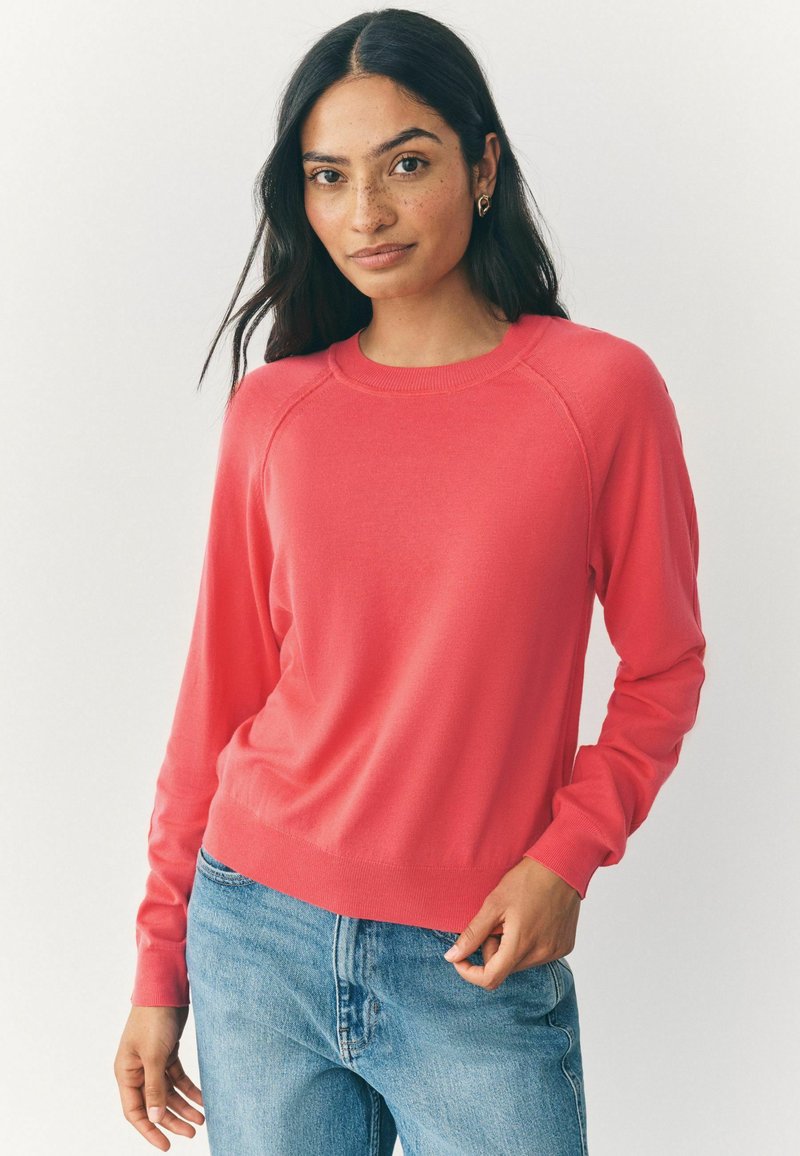 Korallfarbener Strickpullover mit rundem Halsausschnitt, gerippten Bündchen und Saum. Weiche Textur und Raglanärmel, kombiniert mit blauen Jeans.