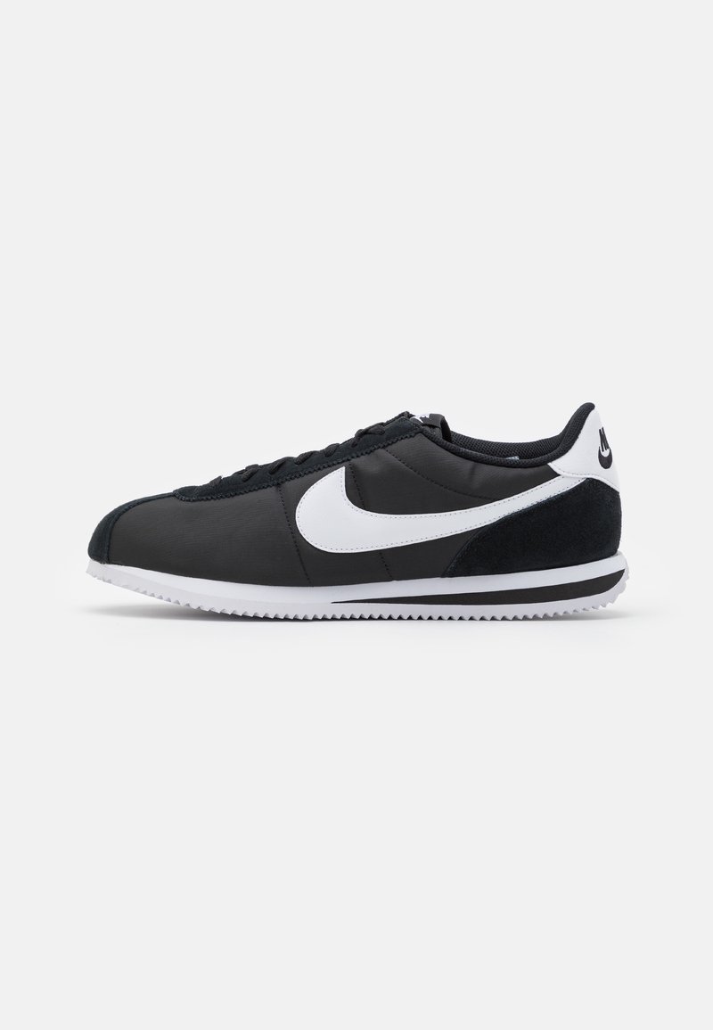 Chaussure de sport noire avec une tige en daim et en synthétique, dotée d'un swoosh Nike blanc, de lacets et d'une semelle en caoutchouc texturé. Accentuée par un talon blanc.