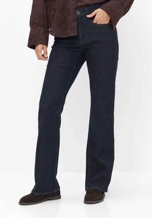Person, die dunkelblaue Straight-Leg-Jeans mit braunen Schuhen und ein braunes, strukturiertes Langarmoberteil trägt, eine Hand in der Tasche.