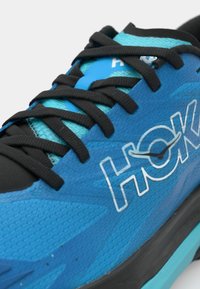 Zapatilla deportiva azul con una superficie texturizada, cordones negros y la marca HOKA. Presenta una parte superior de malla aireada y una suela negra acolchada.