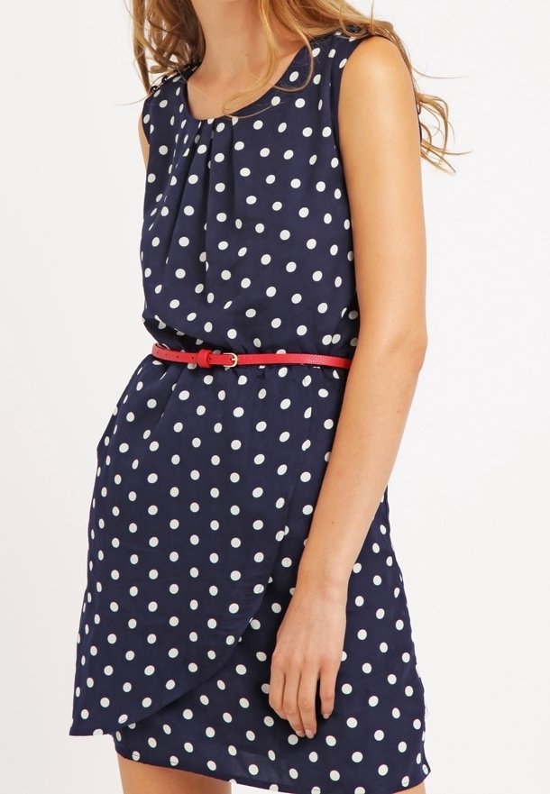 Femme portant une robe bleu marine sans manches à pois blancs et une fine ceinture rouge, debout sur un fond clair uni.