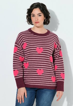 Femme aux cheveux courts et foncés portant un pull rayé violet et rose avec des motifs de cœurs roses et un jean bleu, debout contre un mur blanc.
