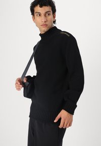 Μαύρο ριμπ πουλόβερ με ψηλό λαιμό και μακριά μανίκια, με μια διακριτική κίτρινη λογότυπο στον ώμο. Κρατιέται με μια μαύρη τσάντα ώμου.