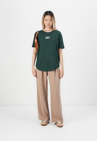Grønn bomulls t-skjorte med hvit "GAP" logo, kombinert med beige vide bukser. Tilleggsbrun skulderveske og mønstrede sko.