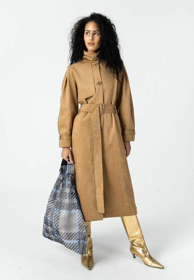 Femme aux cheveux bouclés portant un manteau ceinturé beige et des bottes dorées, tenant un grand sac fourre-tout à motifs bleus et gris, debout devant un fond blanc.