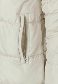 Veste matelassée beige clair avec une poche latérale zippée, présentant des sections capitonn�ées et une texture douce et lisse.