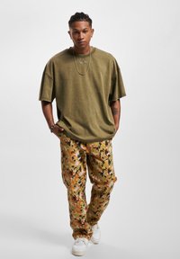 Olive green oversized t-shirt in soft material; tan camo cargo pants with orange and brown patterns; white sneakers; silver chain accessory.