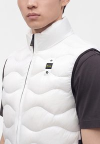 Jeune homme portant un gilet matelassé blanc sans manches avec un col montant par-dessus une chemise noire à manches courtes.