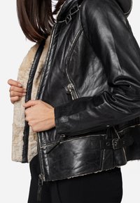 Giacca di pelle nera con interno in shearling, dotata di collo alto, tasche con cerniera e dettaglio con cintura laterale per la regolazione.