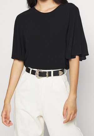 Kobieta ubrana w czarną bluzkę z krótkim rękawem i luźnymi rękawami, białe spodnie z wysokim stanem oraz czarny pasek z ozdobną srebrną klamrą.