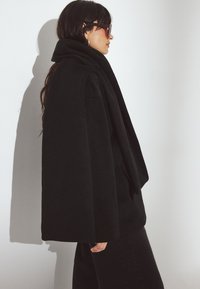 Czarny, oversize płaszcz z miękkiego materiału, z wysokim kołnierzem i drapowanym przodem. Stylizowany z czarnymi dżinsami i okularami przeciwsłonecznymi.