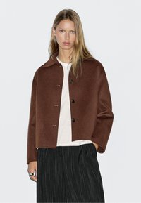 Bruine wolblend cropped jacket met een ronde kraag, voorzien van grote knopen en een rechte silhouet. Draagt over een wit shirt.