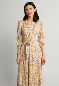 Kwiatowa sukienka typu wrap w miękkim, brzoskwiniowym materiale z niebieskimi i żółtymi wzorami kwiatowymi, dekolt w serek, bufiaste rękawy oraz dopasowany pasek w talii.