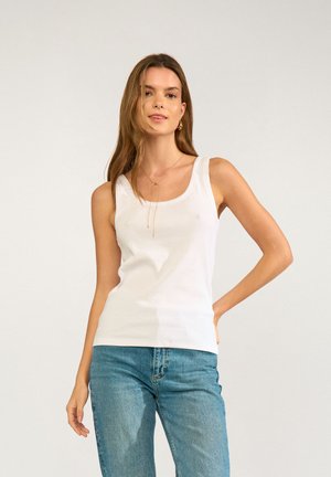 Femme aux longs cheveux bruns portant un haut blanc sans manches, un jean bleu et un délicat collier en or, debout devant un fond uni.