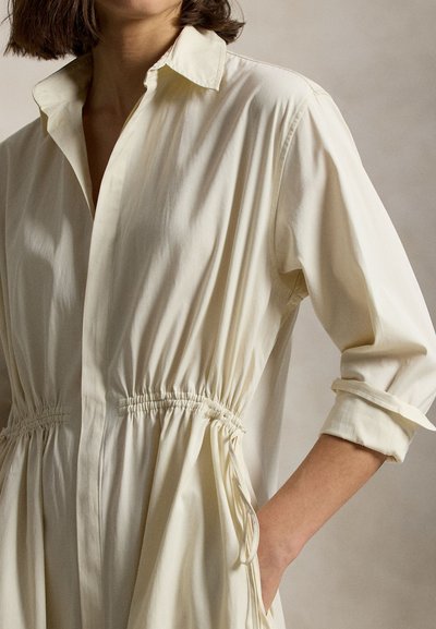 Φόρεμα πουκάμισο σε κρεμ απόχρωση με γιακά, ζώνη με κορδόνι στη μέση και μακριά μανίκια. Ομαλή υφή με άνετη εφαρμογή και πλαϊνές τσέπες.