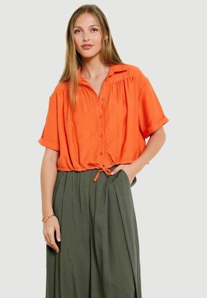 Mladá žena s dlhými blond vlasmi, nosiaca oranžový krátky rukáv so zapínaním na gombíky, so sťahovacím pásom v páse a voľné olivovo zelené nohavice.