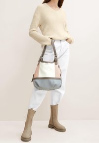 Hobo tas in beige, crème en blauwe tinten, met een zacht materiaal, gebogen vorm en een verstelbare band. Model draagt een gebreide beige trui en witte broek.
