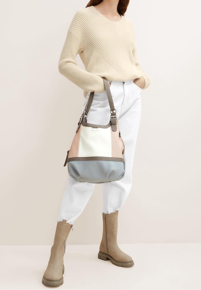 Hobo tas in beige, crème en blauwe tinten, met een zacht materiaal, gebogen vorm en een verstelbare band. Model draagt een gebreide beige trui en witte broek.