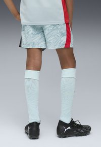 Strój piłkarski składający się z jasno niebieskich spodenek w wzorki, czerwonych akcentów, białych skarpet do kolan o fakturze oraz czarnych korków z zielonymi detalami.