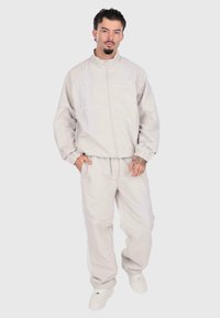 Gaiši pelēks sporta tērps, izgatavots no gludas auduma, ar elastīgiem aprocēm un jostasvietu, ar smalkām baltām akcentiem un logo detaļu uz krūšu un biksēm.