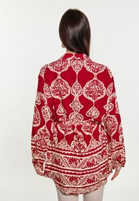 Rød mønstret bluse med indviklede cremede blomster- og geometriske designs, lange ærmer og en detaljeret knyttet talje. Glat stofstruktur.