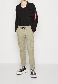 Svart button-up jacka, svart t-shirt, beige cargo byxor med två sidofickor, svart och vita sneakers, och silverkedja.