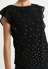 Blusa negra de manga corta con escote redondeado, adornada con lunares dorados, con bordes de mangas con volantes y una textura de tela suave.