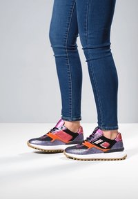 Buty sportowe z połyskującą, fioletowo-różową cholewką, czarnymi akcentami, pomarańczowymi detalami oraz teksturowaną podeszwą z wielokolorowym bieżnikiem.