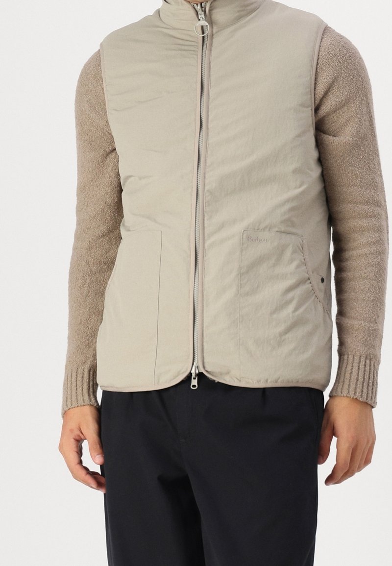 Light beige puffer vest with a high collar, two front pockets, and a zip closure. Features a smooth texture and subtle branding detail.