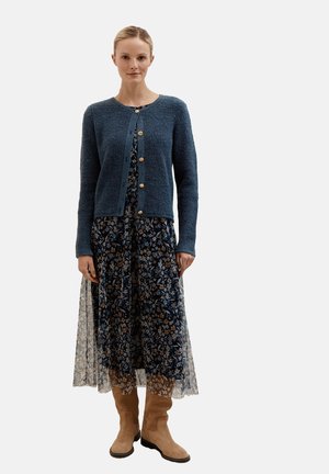 Gilet texturé bleu avec des boutons dorés, superposé sur une robe imprimée florale sombre, associé à des bottes beige jusqu'aux genoux.