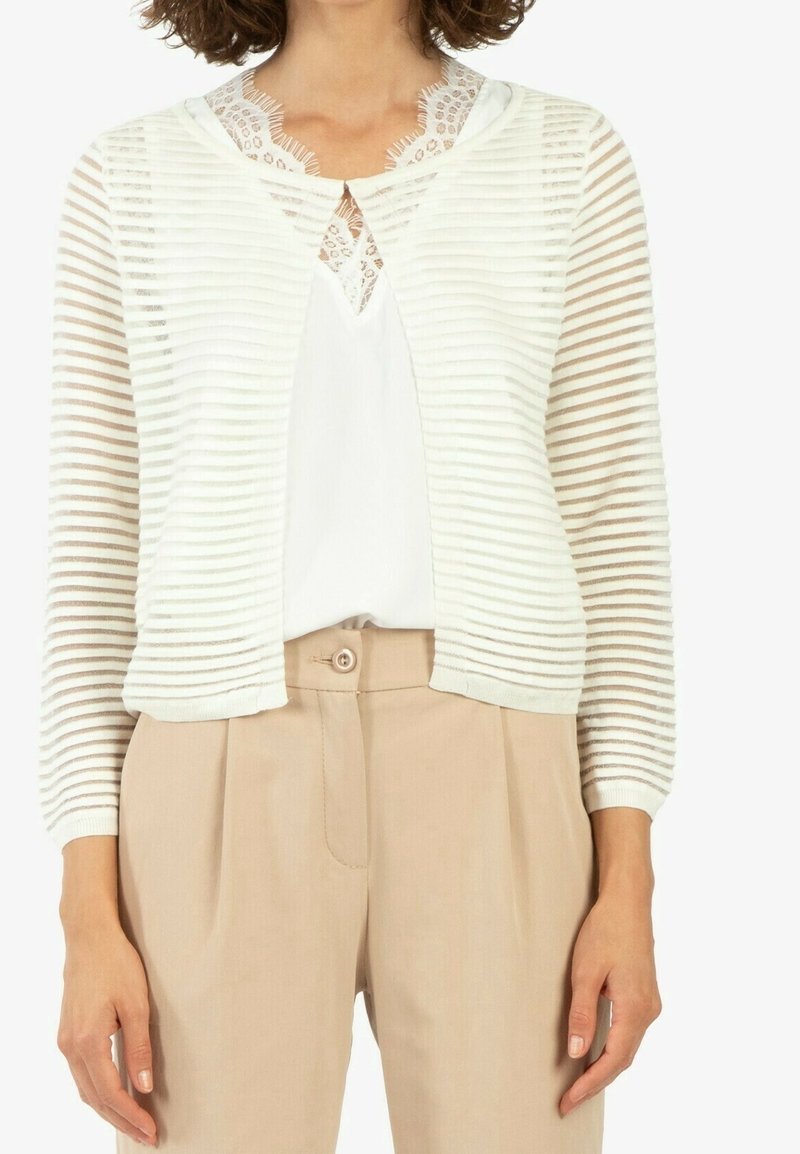 Femme portant un pantalon beige, un haut blanc orné de dentelle, et un cardigan blanc semi-transparent à rayures horizontales et manches trois-quarts.