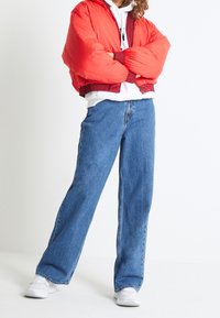 Red padded bomber jacket with hood, ribbed cuffs, and hem, paired with loose-fit blue denim jeans. White sneakers complete the outfit.