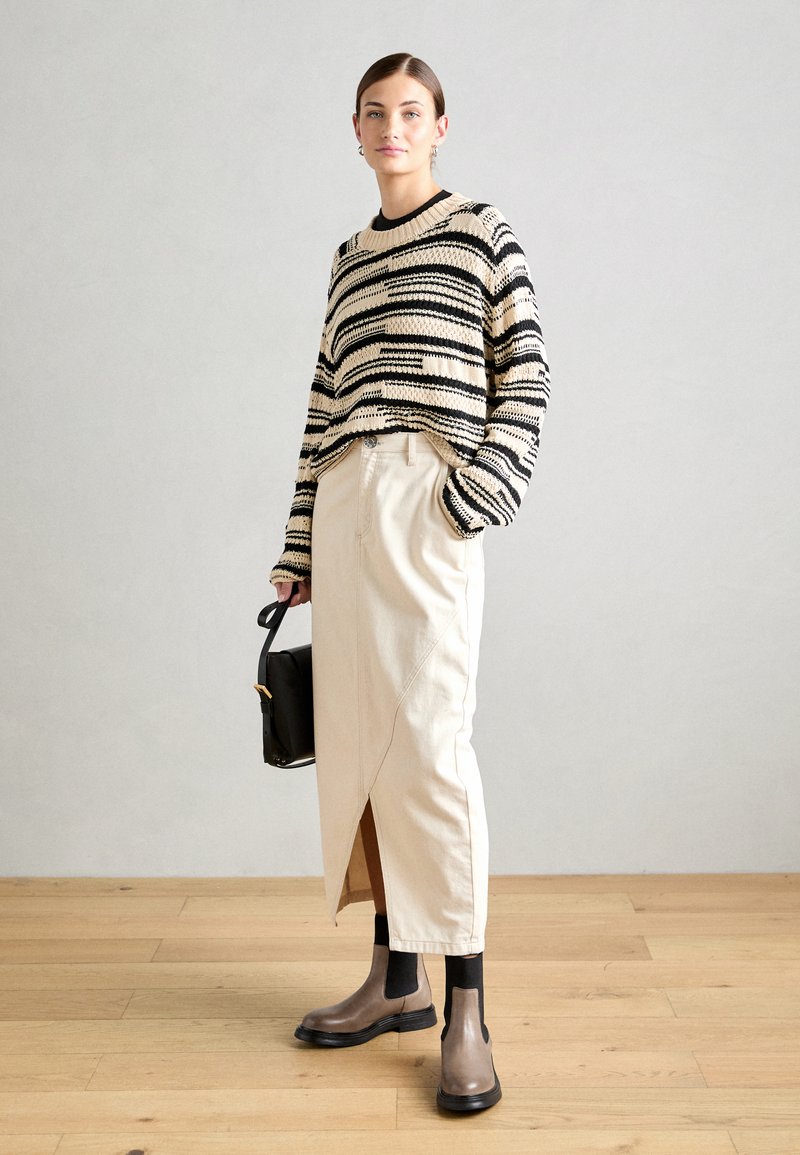 Gestreifter Strickpullover in Creme und Schwarz, beigefarbener Rock mit seitlichem Schlitz, braune Knöchelstiefel und schwarze Handtasche mit Tragegurt. Glatte Texturen, lässiges Design.