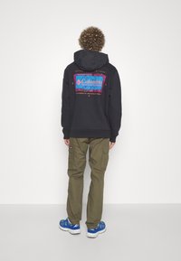 Svart hoodie med en färgglad Columbia-logotyp på ryggen, ihop med olivgröna cargobyxor och blåa träningsskor.