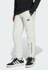 Bijele trenirke s elastičnim pojasom i su�ženim gležnjevima. Imaju crni Adidas logo na lijevoj nozi. Uparene su s crno-bijelim tenisicama.