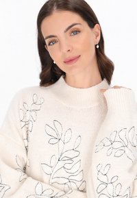 Pulover bej deschis cu guler înalt cu dungi, având broderie florală neagră și accente de mărgele pe mâneci. Țesătură tricotată texturată.