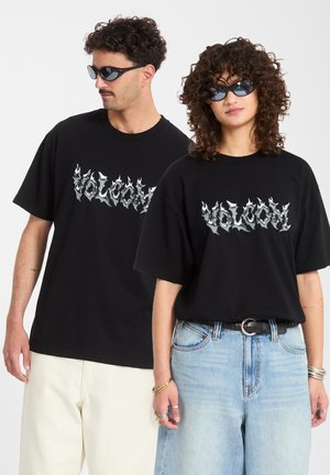 Man en vrouw dragen zwarte Volcom t-shirts met vlammend logo en zonnebrillen; vrouw in lichtblauwe spijkerbroek en accessoires, man in witte broek.
