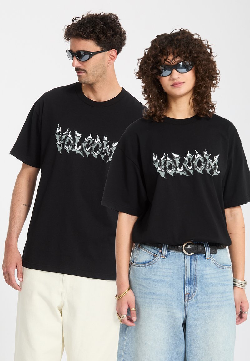 Άνδρας και γυναίκα φορούν μαύρα t-shirts με λογότυπο φλόγας Volcom και γυαλιά ηλίου· η γυναίκα με ανοιχτόχρωμο μπλε τζιν και αξεσουάρ, ο άνδρας με λευκό παντελόνι.