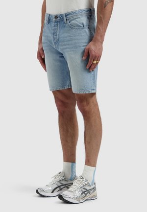 Man draagt lichtblauwe denimkorte broek, witte sokken met blauwe accenten en zilveren hardloopschoenen, staand tegen een effen achtergrond.