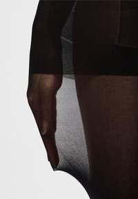 Tessuto nero trasparente con un motivo testurizzato, che mostra una forma liscia e aderente e una leggera traslucenza. Una mano parzialmente visibile sul bordo.