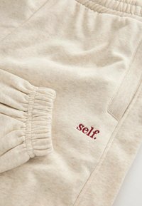 Beżowe spodnie dresowe wykonane z miękkiej tkaniny, z prążkowanym pasem i boczną kieszenią. Na nich wyhaftowane jest słowo „self.” w bordowym kolorze.