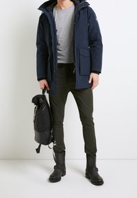Marine parka met capuchon, grijze T-shirt, donkergroene broek en zwarte enkellaarzen; houdt een zwarte trekkingzak met textuur vast.