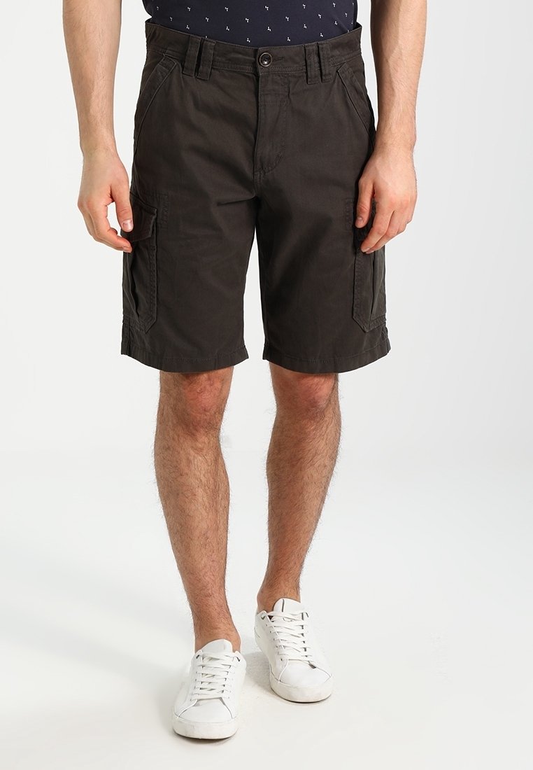 Homme portant un short cargo foncé avec des poches latérales et des baskets blanches à lacets, debout devant un fond blanc uni.