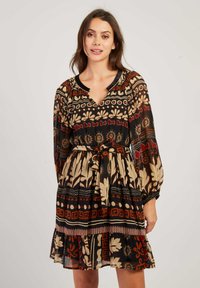 Femme portant une robe longue aux manches longues et arrivant au genou, ornée de motifs tribaux multicolores et dotée d'une ceinture nouée, se tenant devant un fond uni.