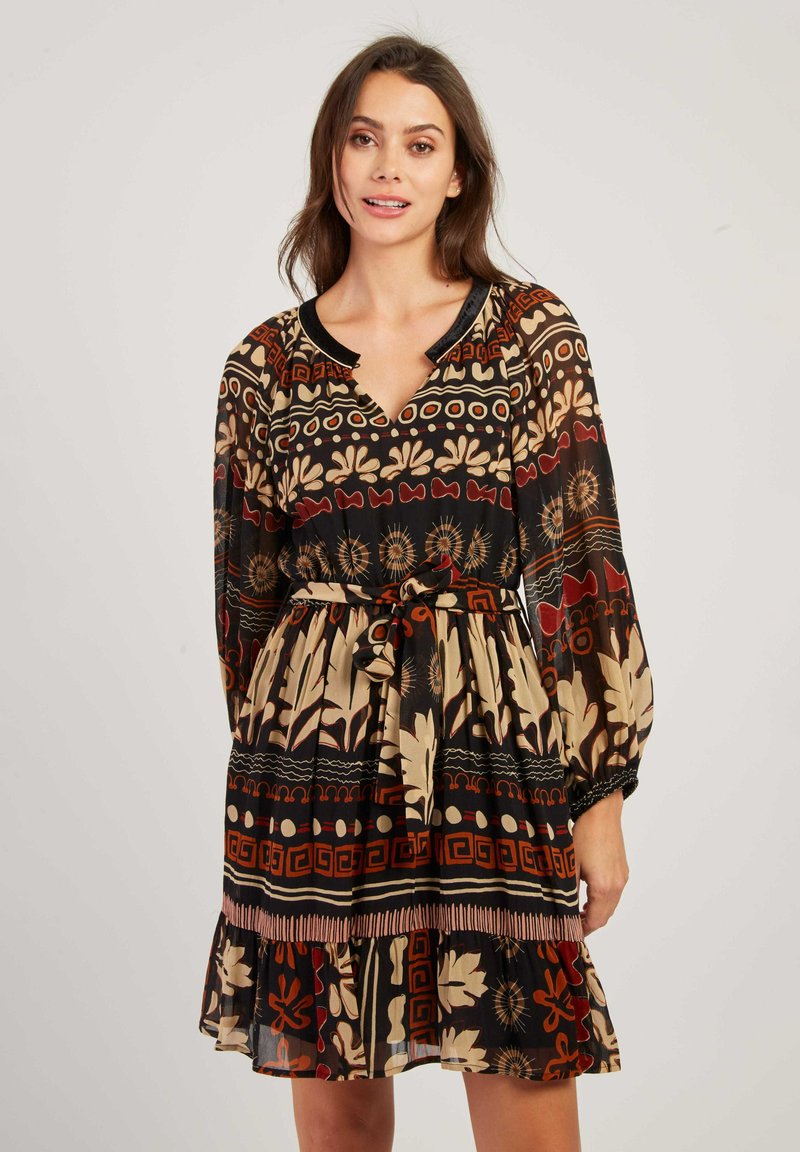 Femme portant une robe longue aux manches longues et arrivant au genou, ornée de motifs tribaux multicolores et dotée d'une ceinture nouée, se tenant devant un fond uni.