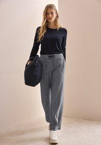 Maglietta a maniche lunghe blu navy, pantaloni grigi larghi con cordoncino e sneaker bianche. Il modello tiene una borsa nera. Texture morbide e design semplice.