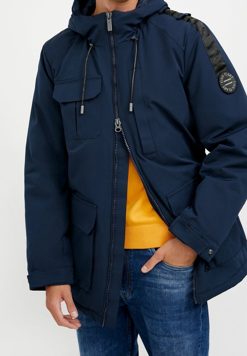 Tmavě modrá parka se zipem vpředu, kapucí na šňůrku a dvěma velkými předními kapsami. Má černé ramenní detaily a žlutou vnitřní vrstvu.