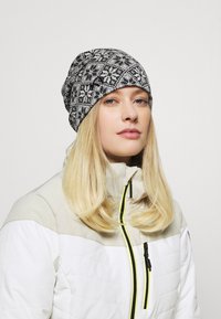 Grå och svart mönstrad mössa i en slouchy design. Bärs med en vit isolerad jacka med svarta och gula accenter.