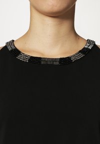 Svart topp med rund halsringning prydd med en detaljerad pärlkrage i olika svarta och grå nyanser, som framhäver olika texturer.