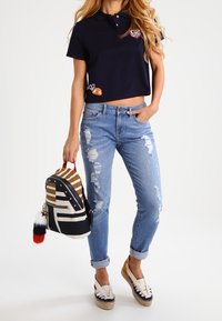 Camisa polo marina con parches bordados, vaqueros claros desgastados, mochila a rayas con asa roja y zapatos de esparto.